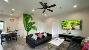 A modern living room with a black sofa, a coffee table, and a ceiling fan. There is a dining area with a table and chairs, and a TV mounted on the wall.