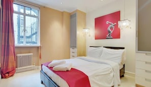 Habitación de hotel con una cama, un radiador, un ventanal con cortina, una pared con un cuadro rojo y una araña.