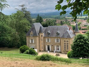 Un grand bâtiment en pierre, doté de nombreuses cheminées, entouré de verdure et d’une zone délimitée par une clôture.