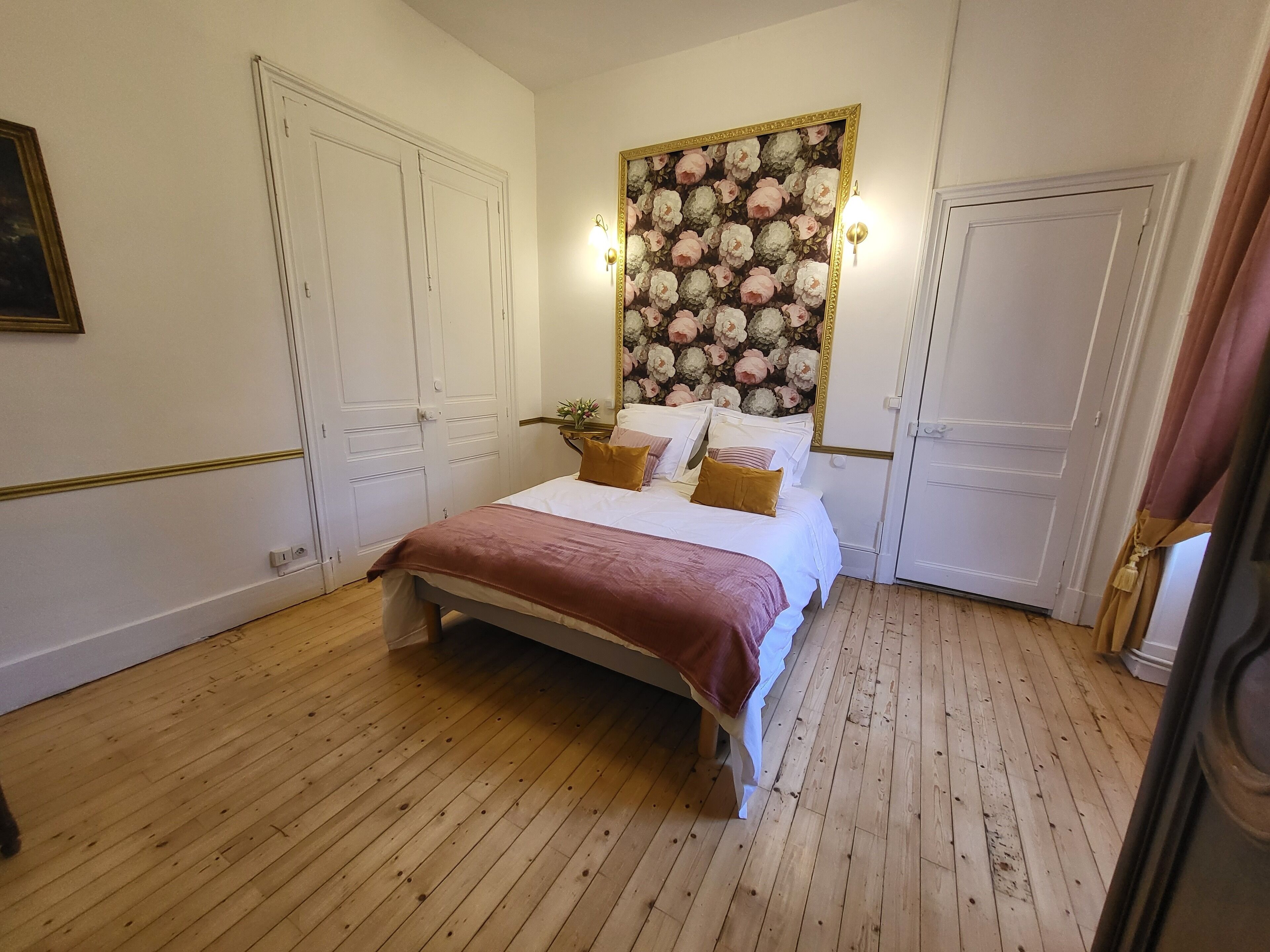 Une chambre avec un plancher en bois, un lit avec une literie blanche et un plaid marron, une tête de lit recouverte d’un papier peint à motifs floraux et des portes blanches.
