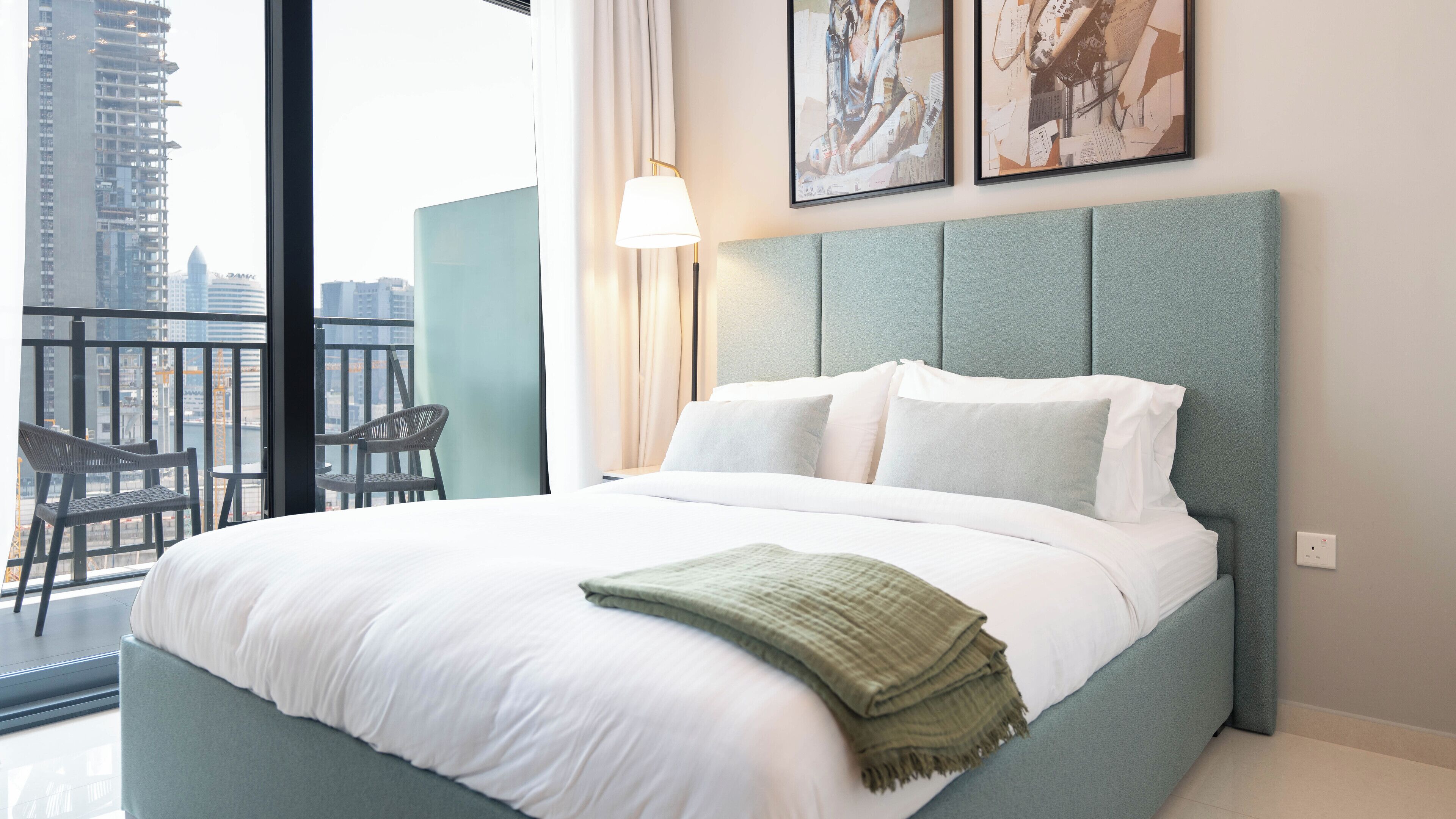 A hotel room with a bed, a chair, a lamp, and a view of the cityscape.