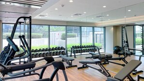 Ein Fitnessstudio mit Trainingsgeräten, Gewichtsständern und großen Fenstern mit Blick auf Grünflächen und Gebäude.