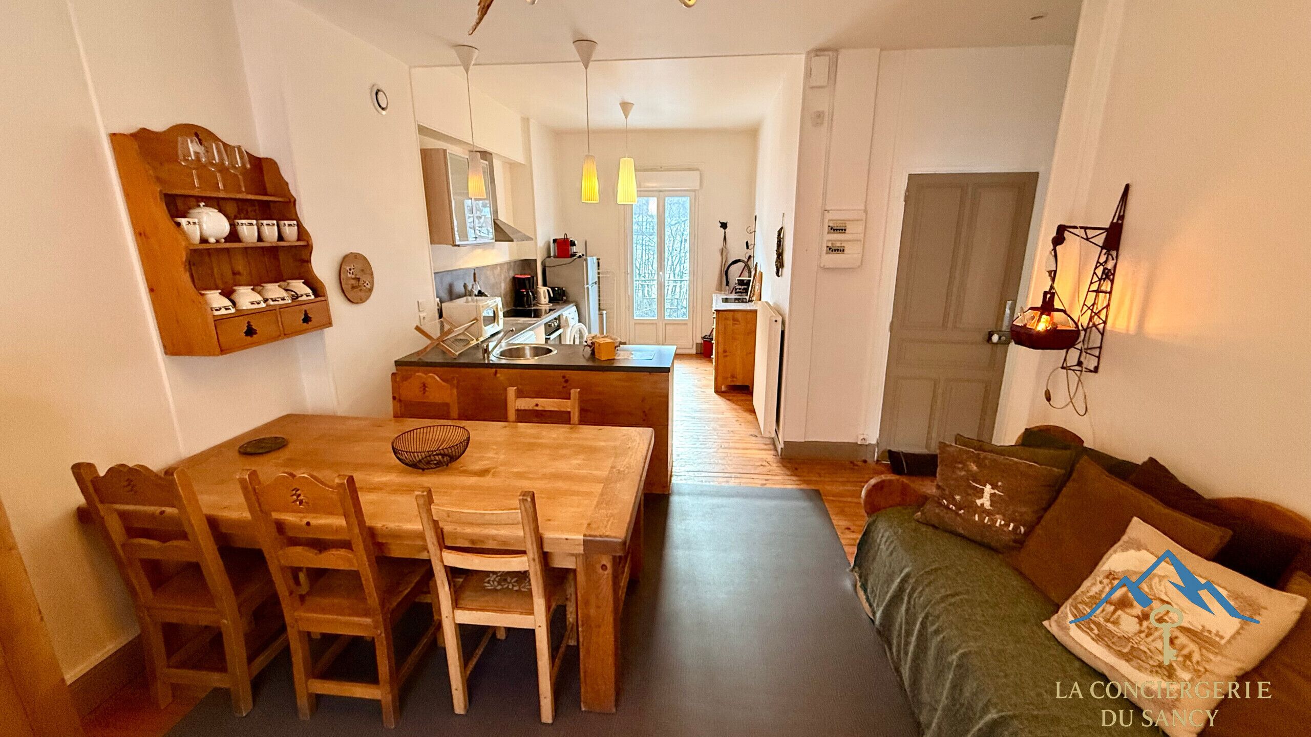 A cozy living room with a wooden dining table, chairs, and a sofa. A kitchen with a sink and stove is visible in the background.