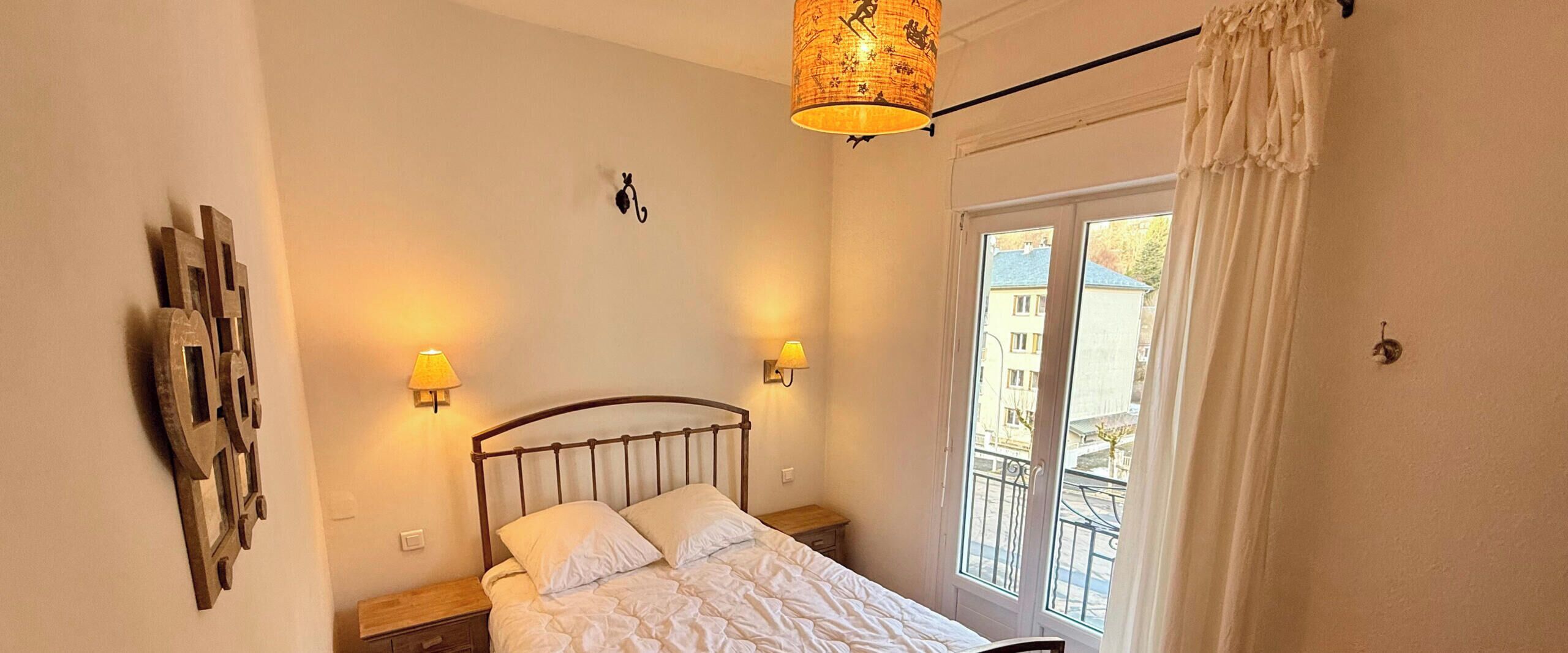A bedroom with a bed, two wall-mounted lamps, a ceiling light, a window with a view of the outside, and a curtain.
