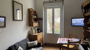 Een gezellige woonkamer met een bank, een eettafel met geruit tafelkleed, een televisie en een raam met uitzicht op bomen.