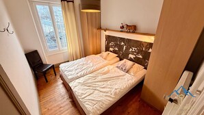 Un dormitorio con dos camas, una silla, un ventanal con cortinas y un estante con una decoración navideña de Papá Noel y vaca.