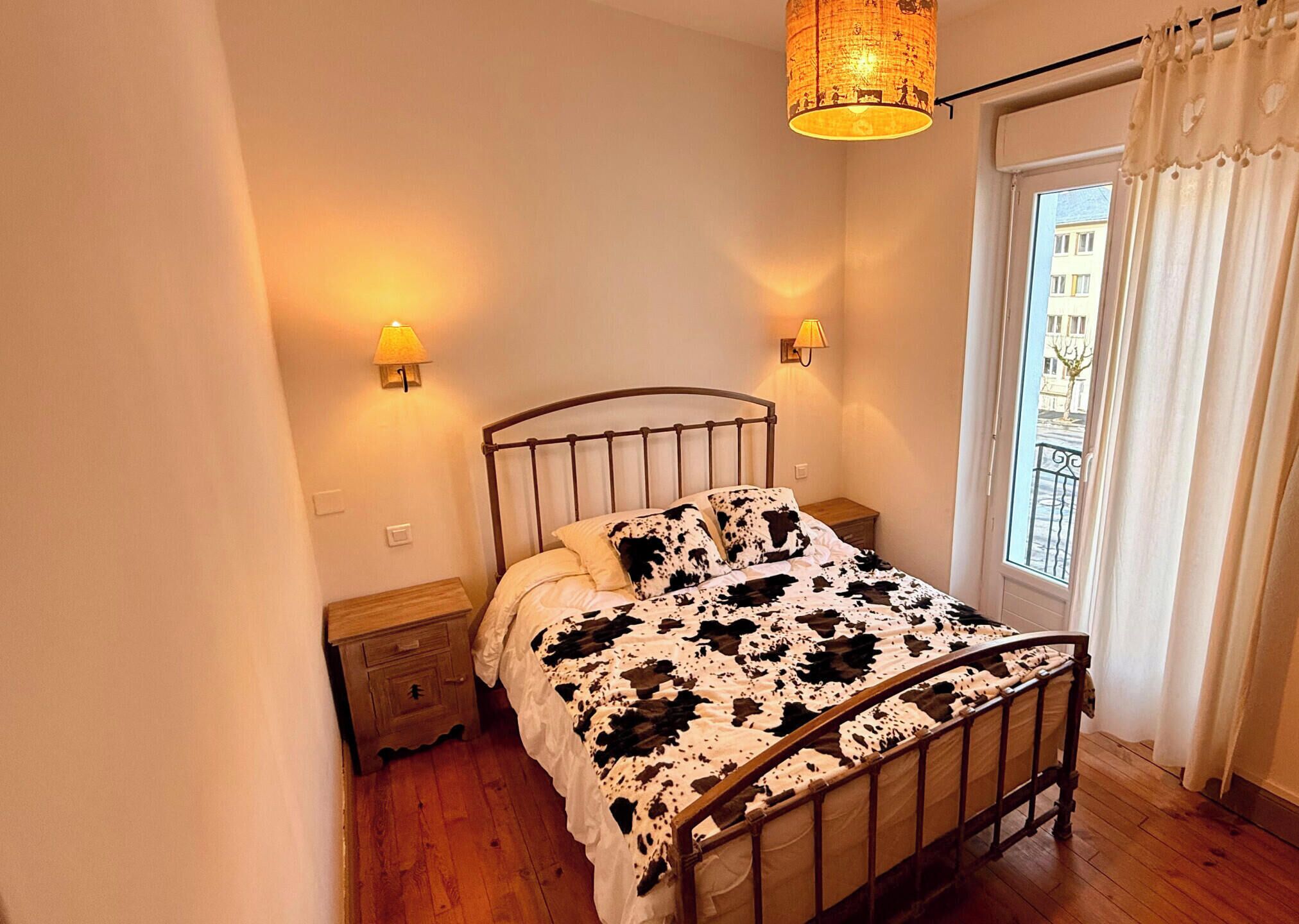 Een slaapkamer met een metalen bedframe, een houten nachtkastje, twee wandlampen, een raam met gordijnen en een hanglamp.