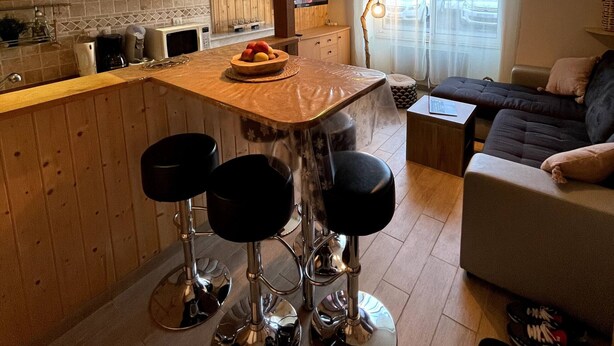 A modern kitchen with wooden cabinets, a dining area with bar stools, a television, and a large window with curtains. - La Féessence boisée - 2/4 pers (Mont-Dore)