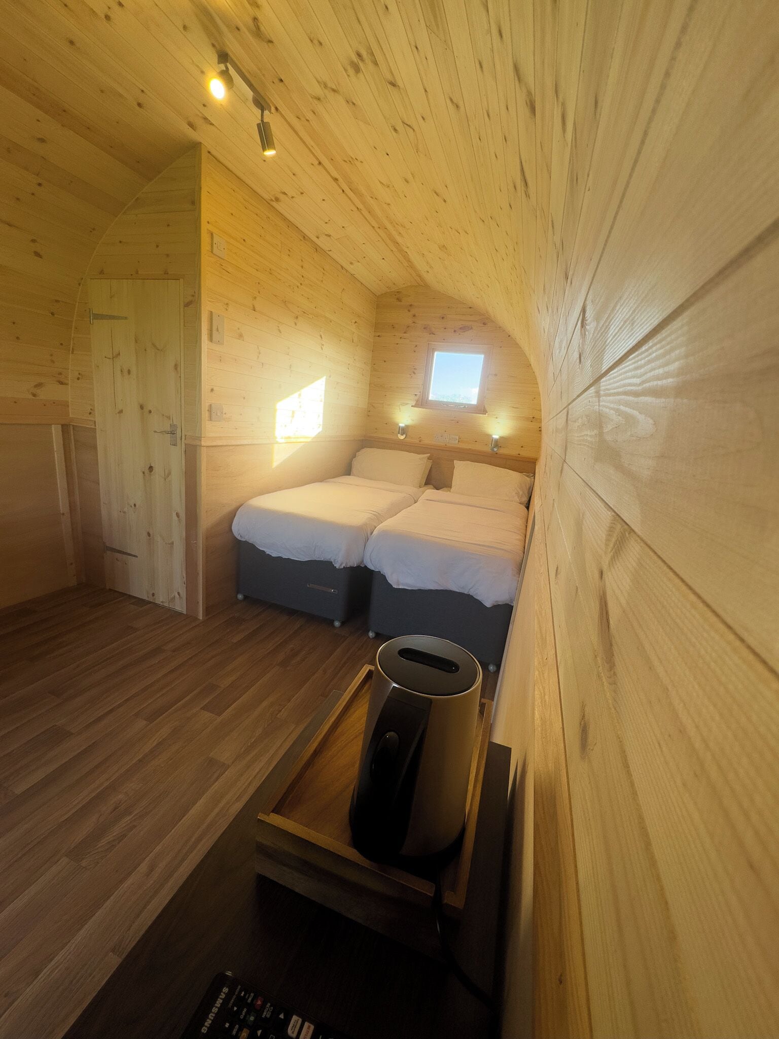 A cozy cabin room with wooden walls and a sloped ceiling. There are two beds with white bedding, a small window, and a ceiling light.