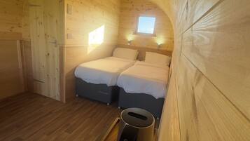 A cozy cabin room with wooden walls and a sloped ceiling. There are two beds with white bedding, a small window, and a ceiling light.