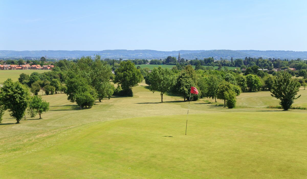 Un campo de golf con un mástil de bandera al frente, rodeado de árboles y zonas verdes abiertas.