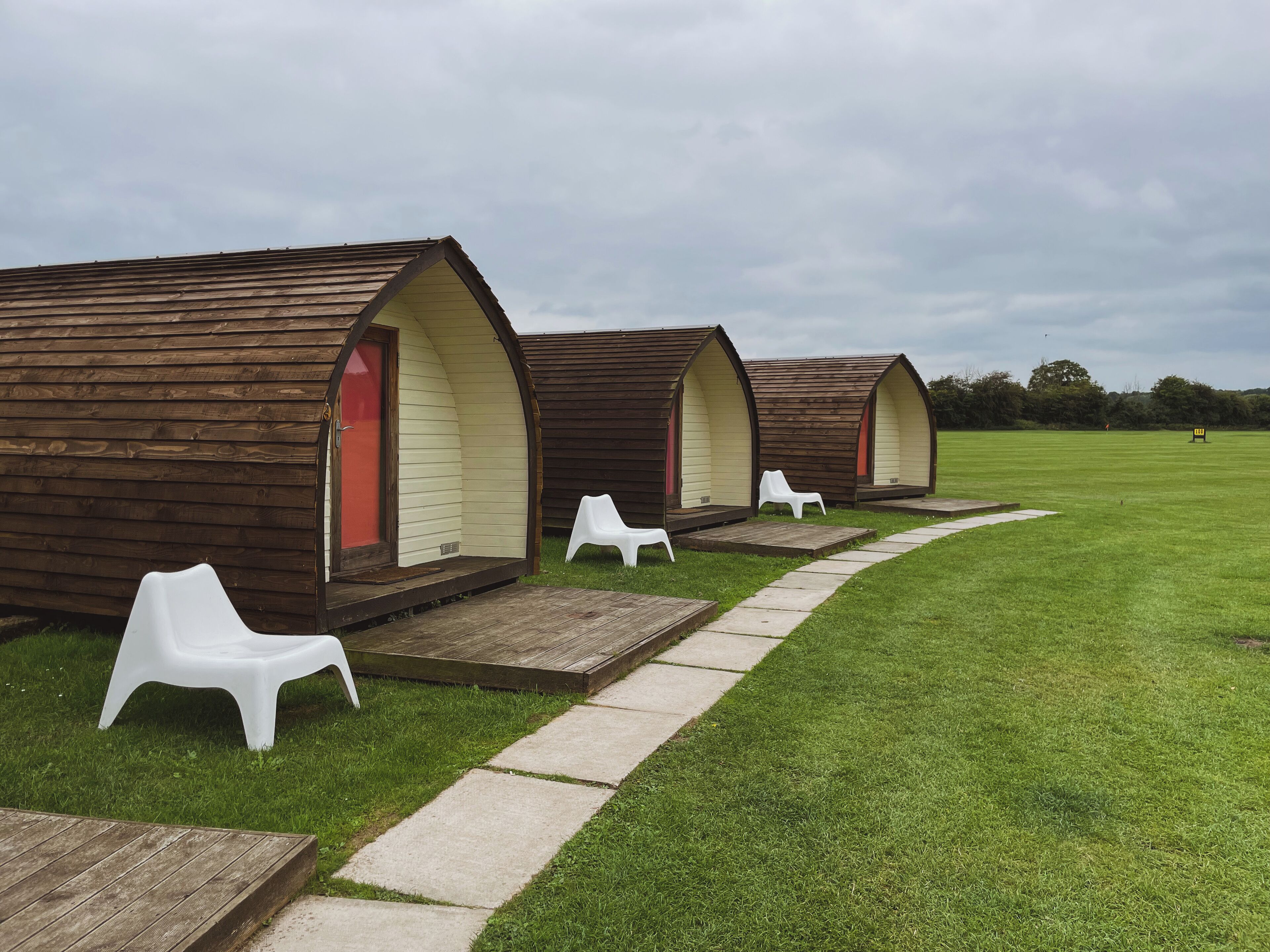 A row of wooden A-frame cabins with white plastic chairs outside, set on a grassy field.