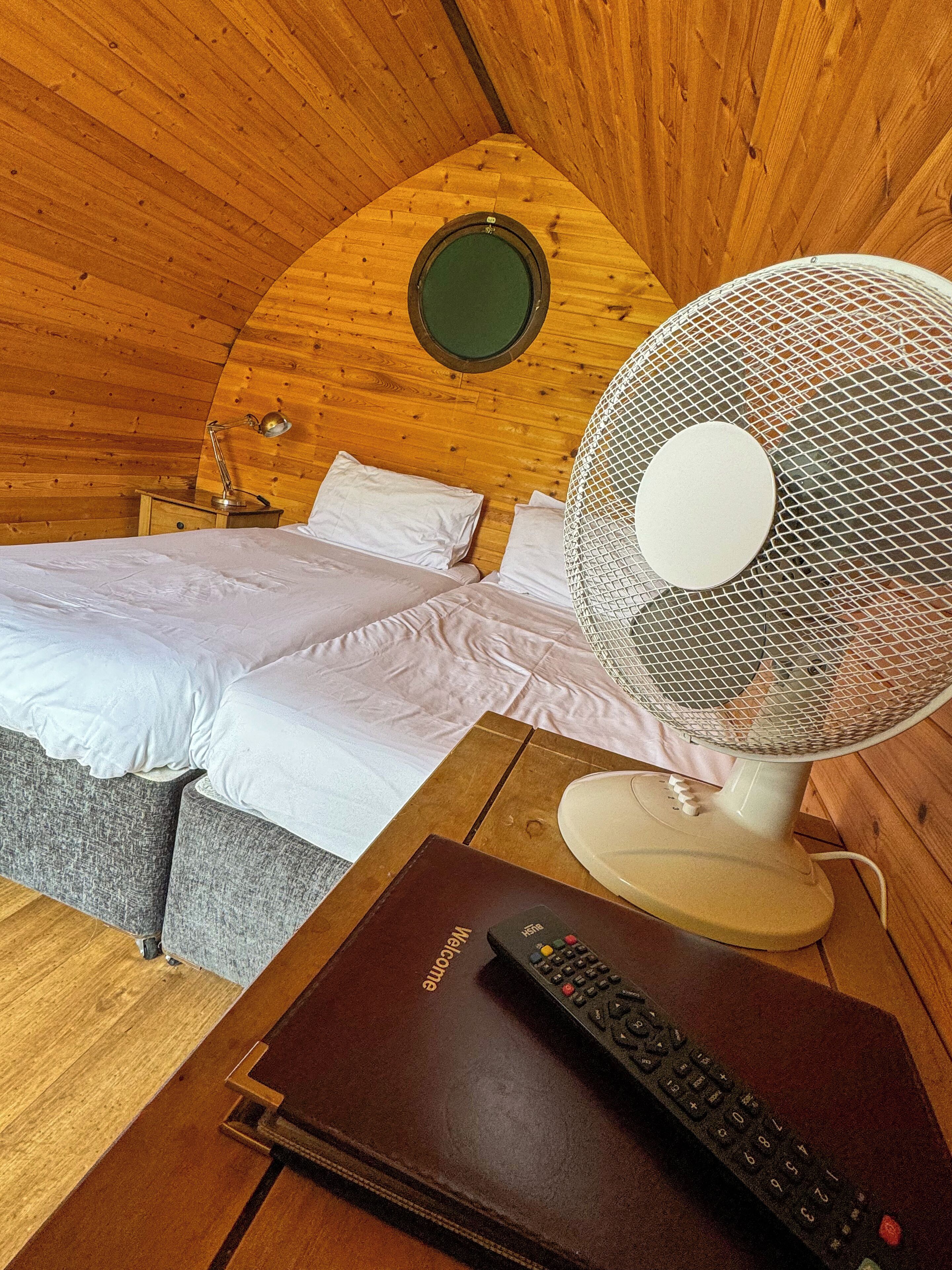 A cozy room with a bed, a standing fan, a remote control, and a wooden ceiling.