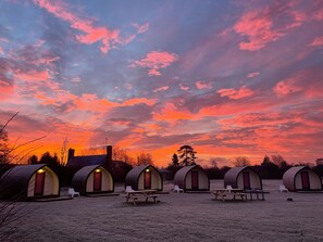 A row of small, dome-shaped cabins with picnic tables in front, set against a sunset sky. - Rodway Hill Eco Pod Hotel (Gloucester)