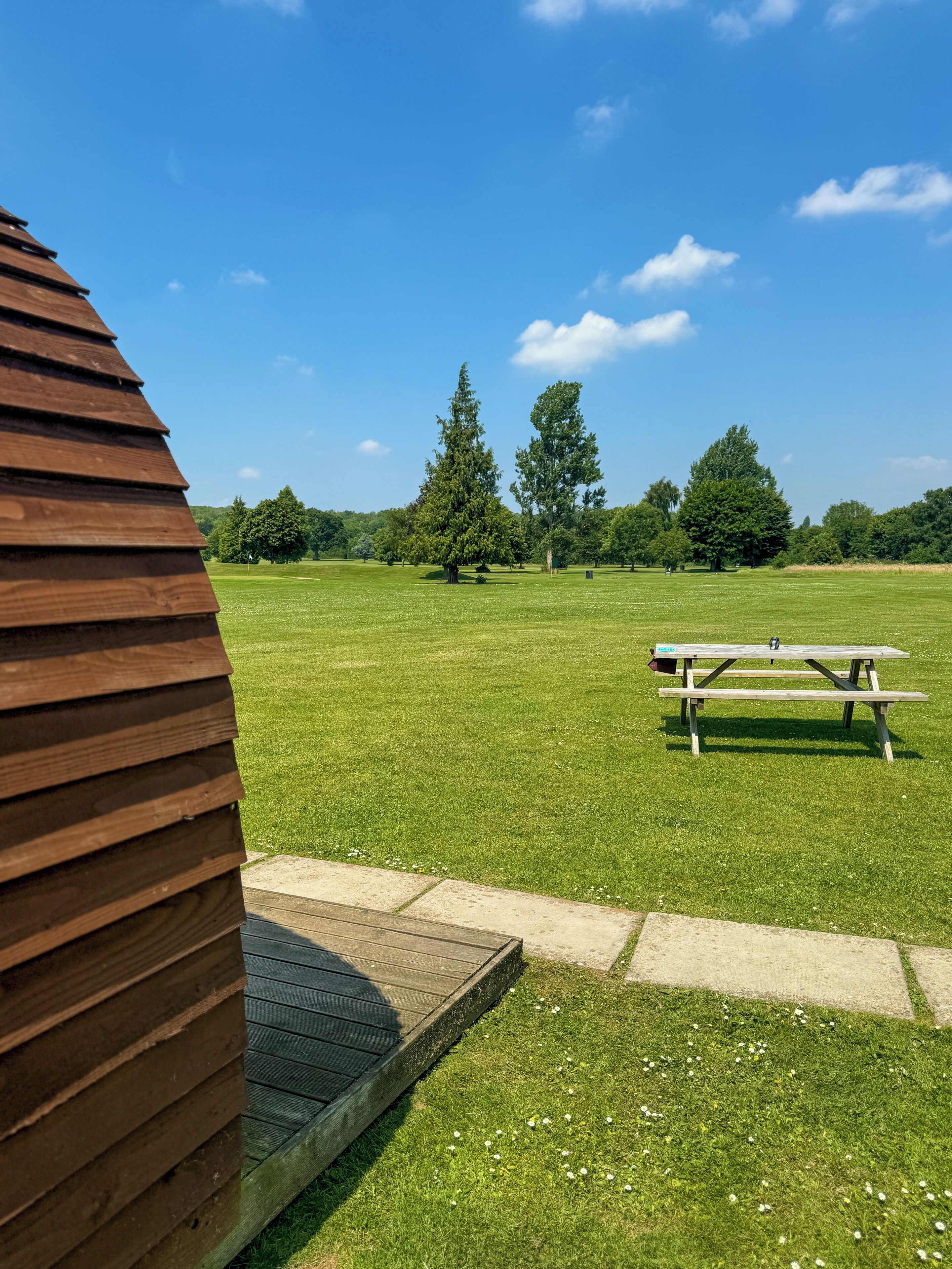 A wooden deck leading to a picnic table in a grassy field with trees in the background.