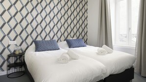 Een hotelkamer met een modern hoofdeinde met een geometrisch patroon, een opgemaakt bed met witte lakens en twee blauwe kussens, een klein nachtkastje met een lamp, en een raam met gordijnen.