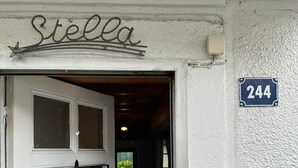 Eine weiße Tür mit einem schwarzen Metallgitter, ein Schild mit der Aufschrift "Stella" darüber und ein blaues Hausnummernschild "244" an der Wand.