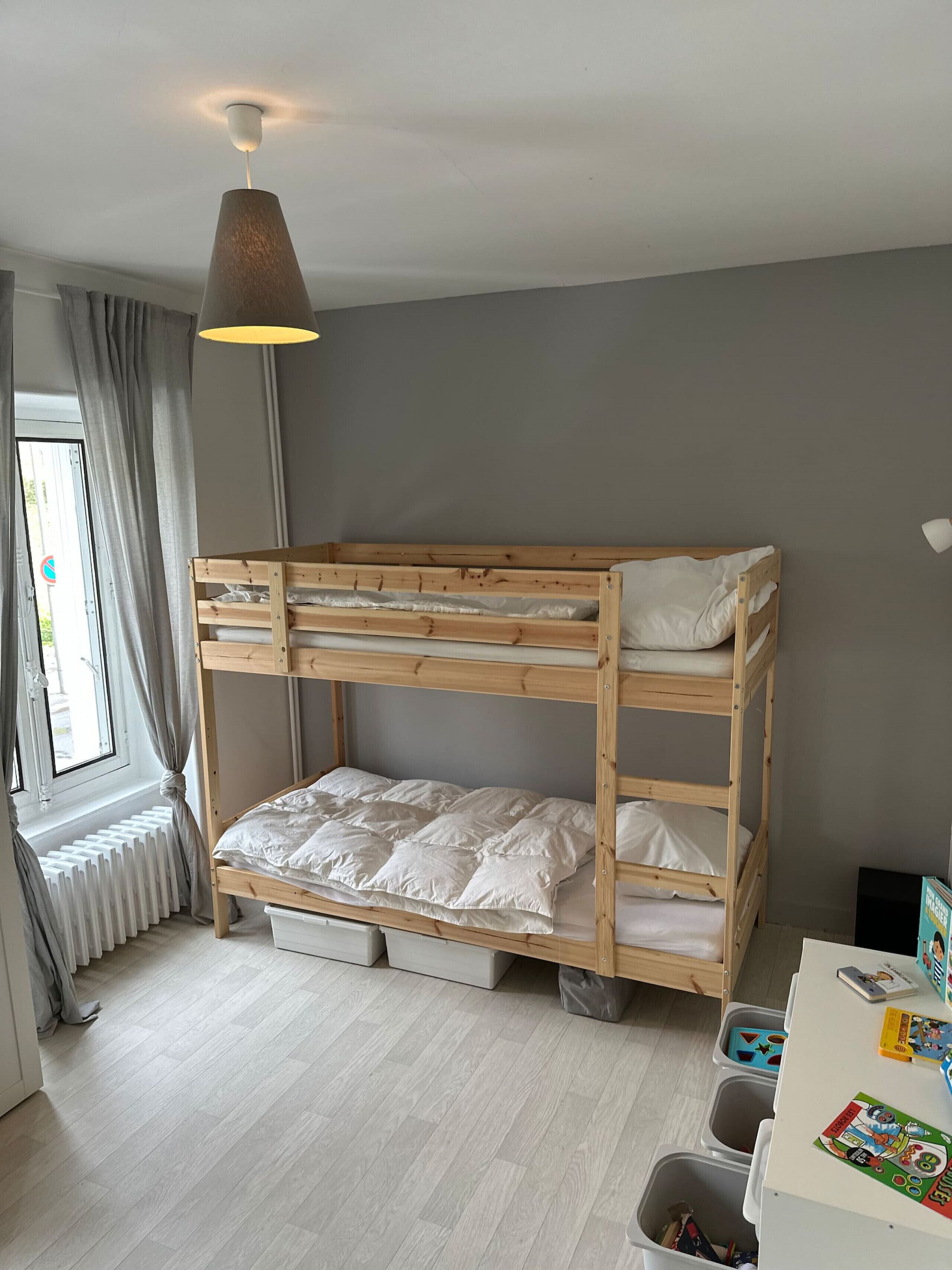 Een stapelbed met opbergladen, een radiator, een raam met gordijnen en een tafel met kinderboeken.