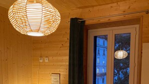 Una habitación acogedora con paredes de madera, una cama con ropa de cama blanca, una mesita de noche con una lámpara y una ventana con vistas a edificios.