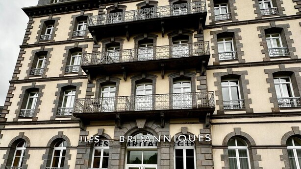 Un immeuble à plusieurs étages de style architectural classique, doté de balcons ornés de rampes en fer forgé et d’une enseigne sur laquelle on peut lire « LES BRITANNIQUES ».