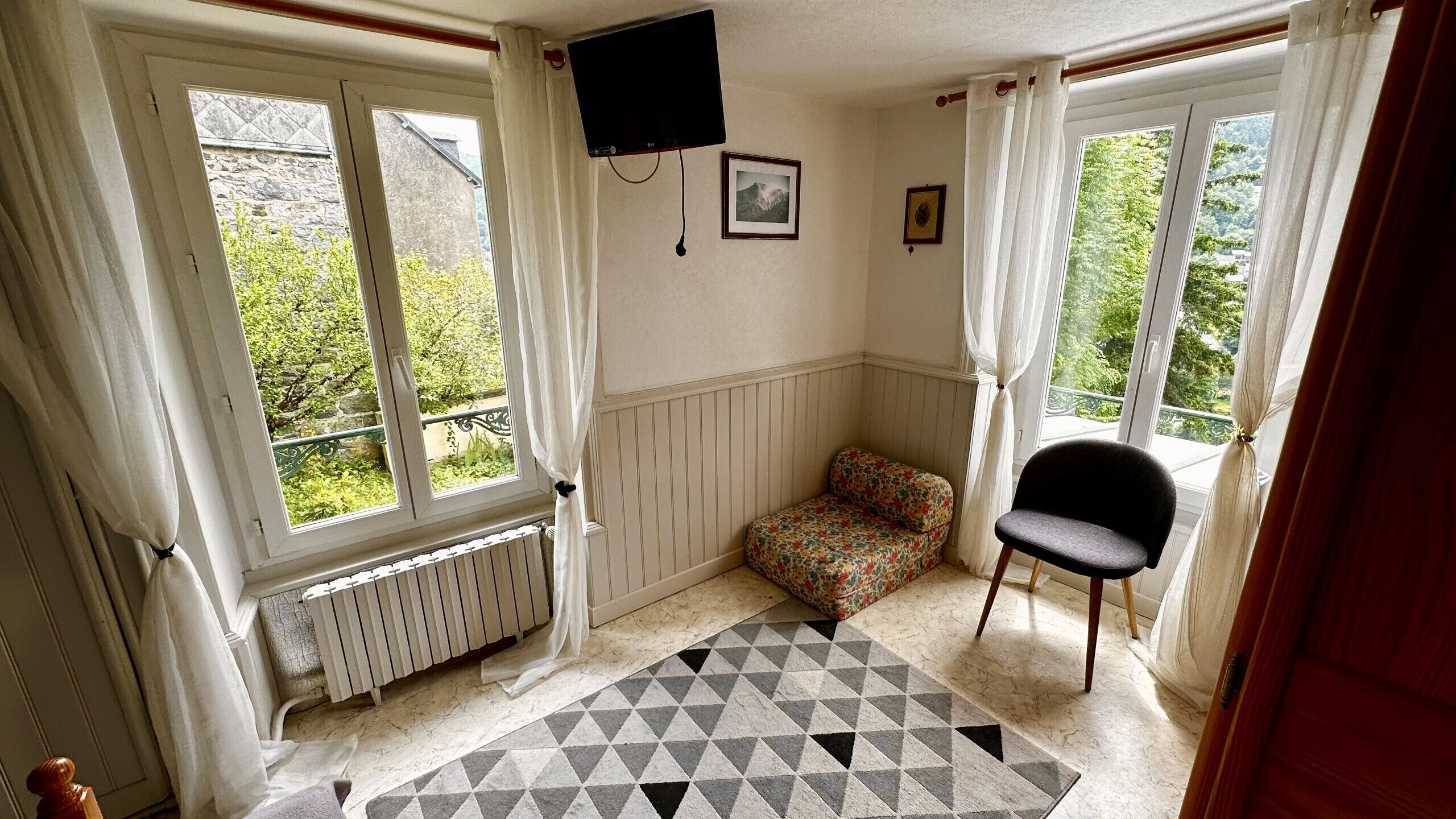 Ein kleiner, gemütlicher Raum mit Heizkörper, einer blumigen Couch, einem schwarzen Sessel und einem an der Wand befestigten Fernseher.