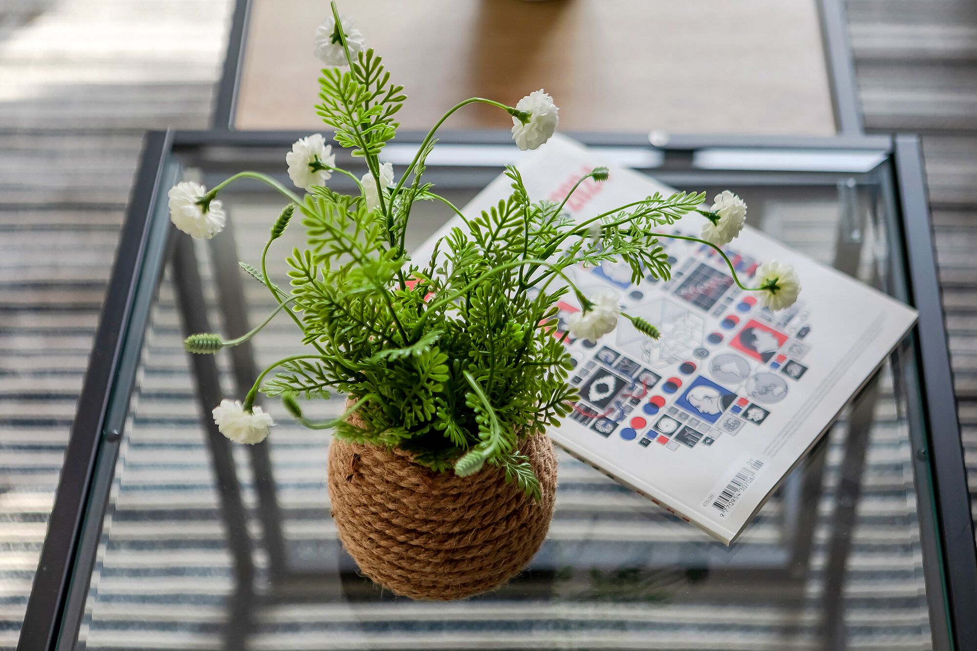 A glass table with a woven basket containing white flowers and green foliage. A magazine is placed on the table.