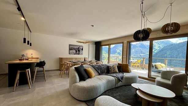 Un salon moderne doté d’un grand canapé d’angle gris, d’un coin repas avec des chaises en bois et d’une vue sur les montagnes par de grandes fenêtres.