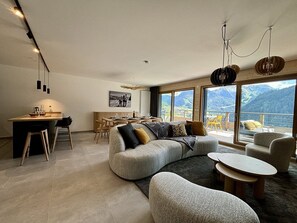 A modern living room with a large gray sectional sofa, a dining area with wooden chairs, and a view of mountains through large windows.