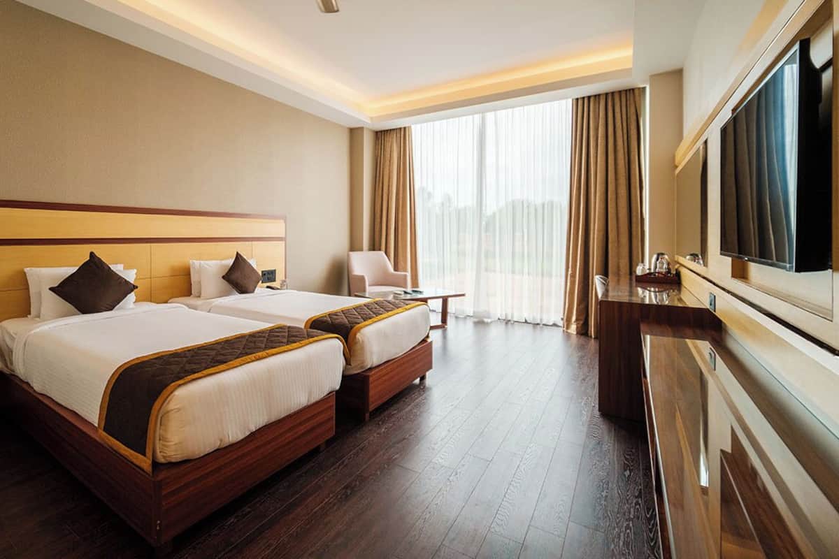 A hotel room with two beds, a TV, a desk, and a large window with curtains.