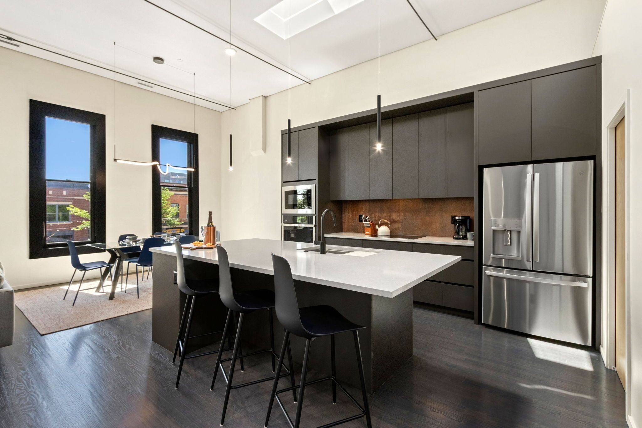 Une cuisine moderne dotée d’un grand îlot central, de meubles de rangement aux couleurs sombres, d’appareils électroménagers en acier inoxydable et offrant une vue sur l’extérieur.