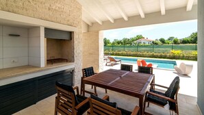 An outdoor patio with a stone wall, a wooden table, and chairs, overlooking a swimming pool and greenery. - Villa Jan in Valbandon (Valbandon)