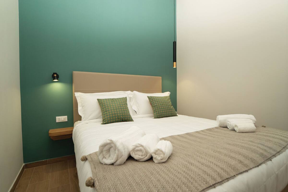 Un letto rifatto con lenzuola bianche e cuscini fantasia verde, una coperta beige e asciugamani arrotolati su un comodino in legno.