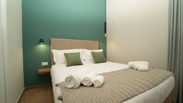 Una cama bien tendida con sábanas blancas y almohadas estampadas en verde, una manta color beige y toallas enrolladas sobre una mesita de noche de madera.