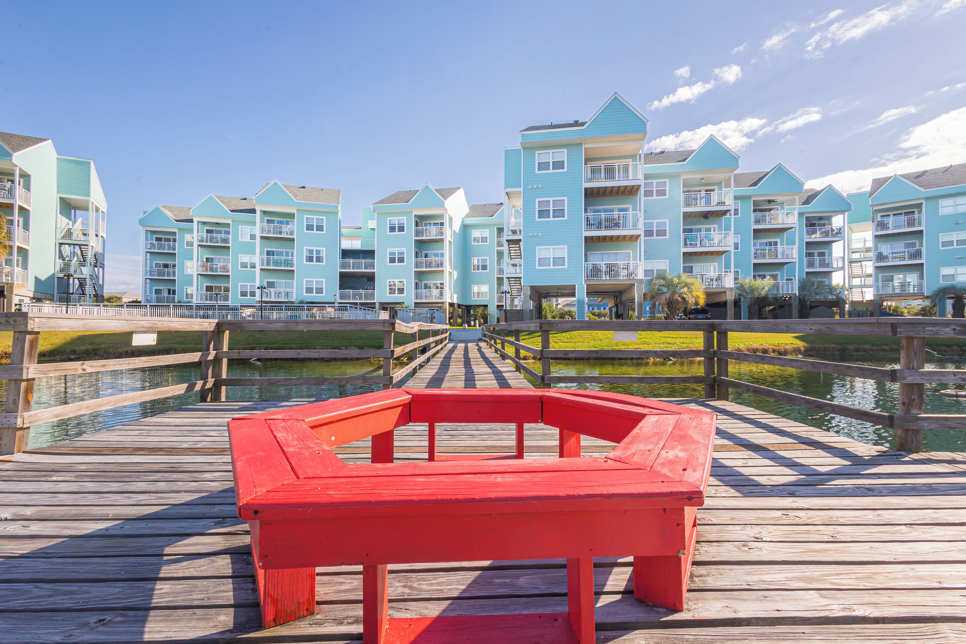 Une jetée en bois mène à une rangée de bâtiments bleu clair dotés de balcons. Un banc rouge se trouve au premier plan, et une étendue d’eau calme se profile à l’arrière-plan.