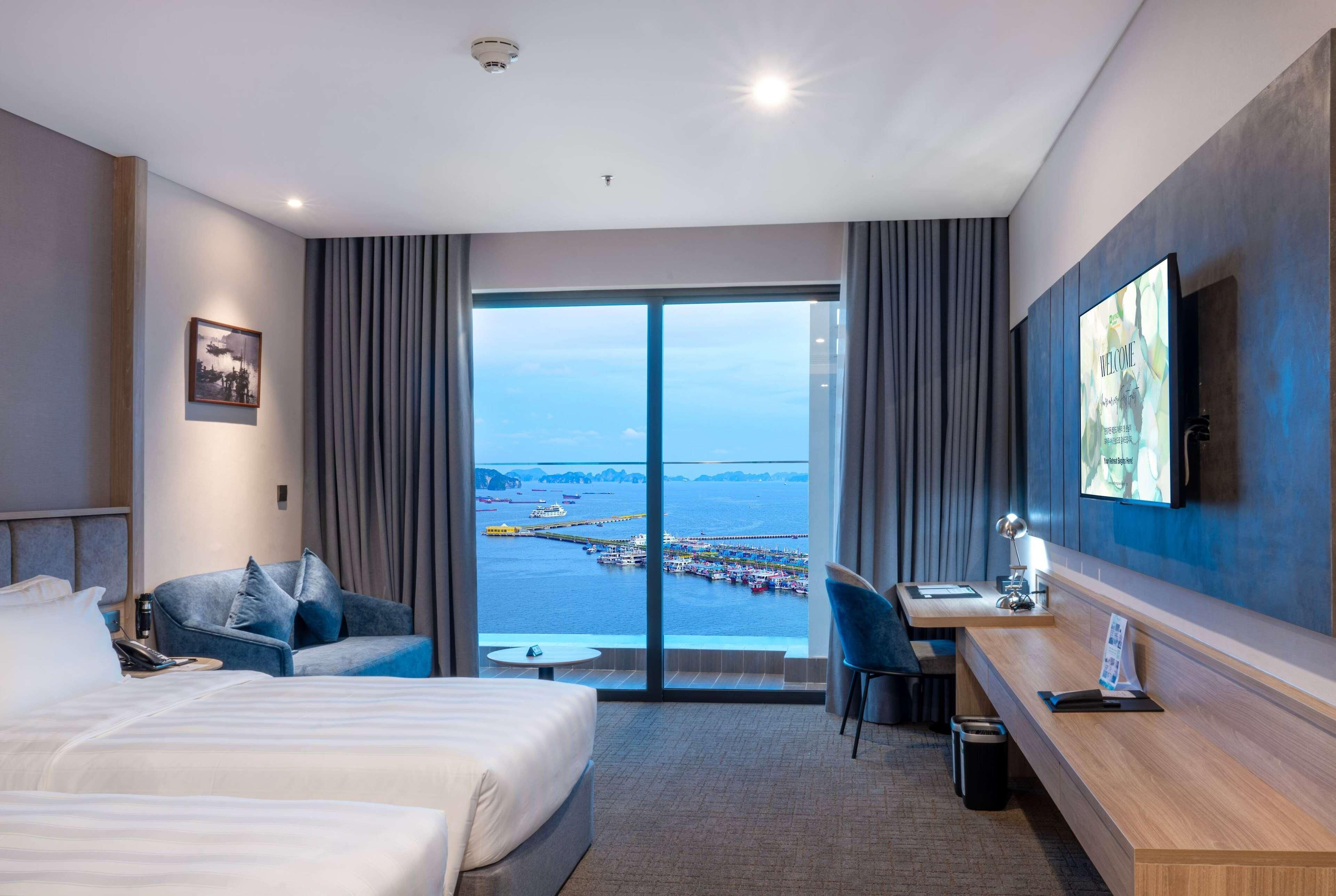 A hotel room with a bed, a desk, a chair, a TV, and a large window with a view of the sea and a pier.