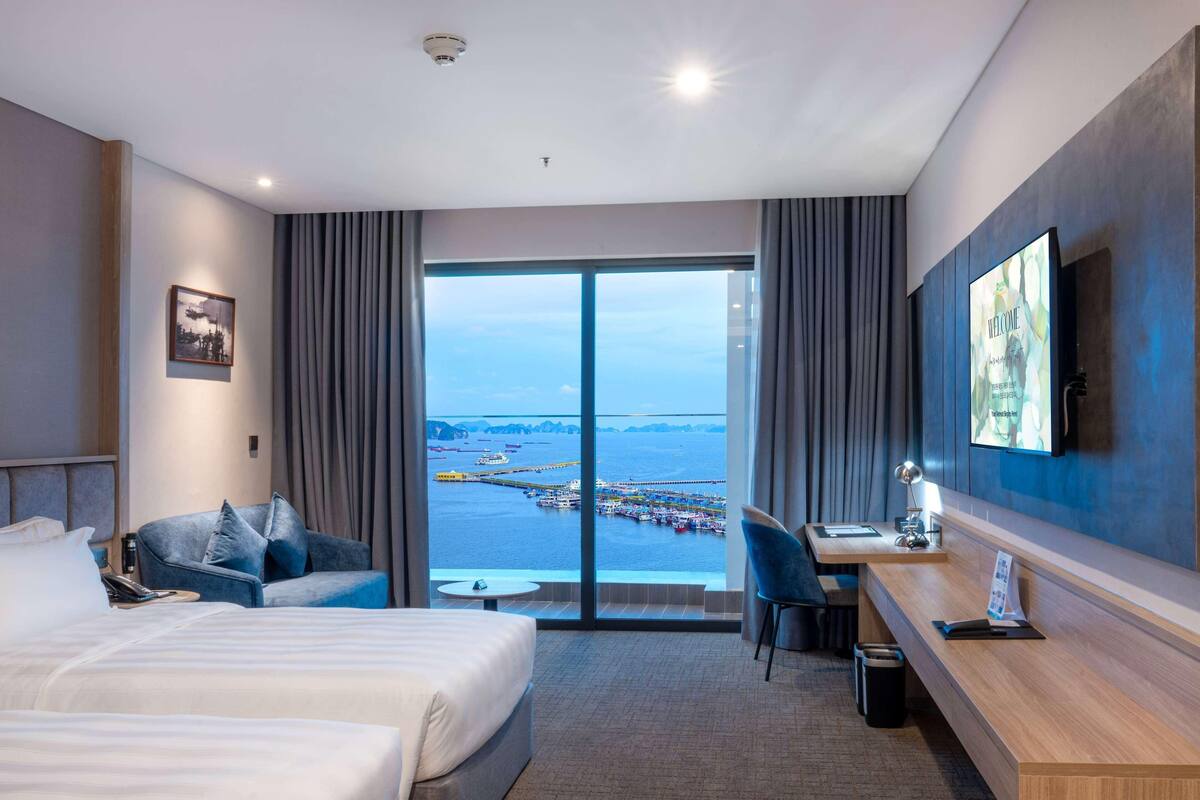 A hotel room with a bed, a desk, a chair, a TV, and a large window with a view of the sea and a pier.