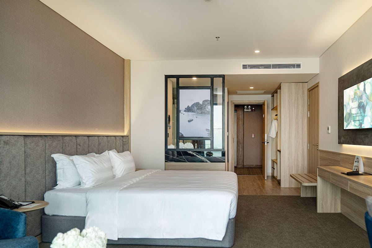 A hotel room with a bed, a desk, a TV, and a view of the sea.