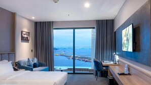 A hotel room with a bed, a desk, a chair, a TV, and a large window with a view of the sea and a pier. - Wyndham Garden Legend Halong (Bai Chay)