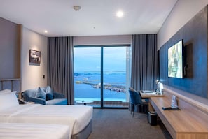 Une chambre d’hôtel équipée d’un lit, d’un bureau, d’une chaise, d’une télévision et d’une grande fenêtre donnant sur la mer et une jetée.