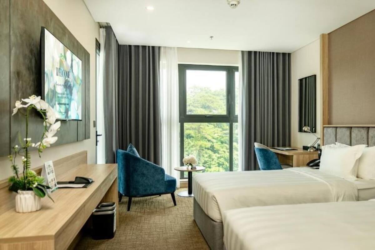 A hotel room with two beds, a desk, a chair, a small table with a vase, and a large window with curtains.