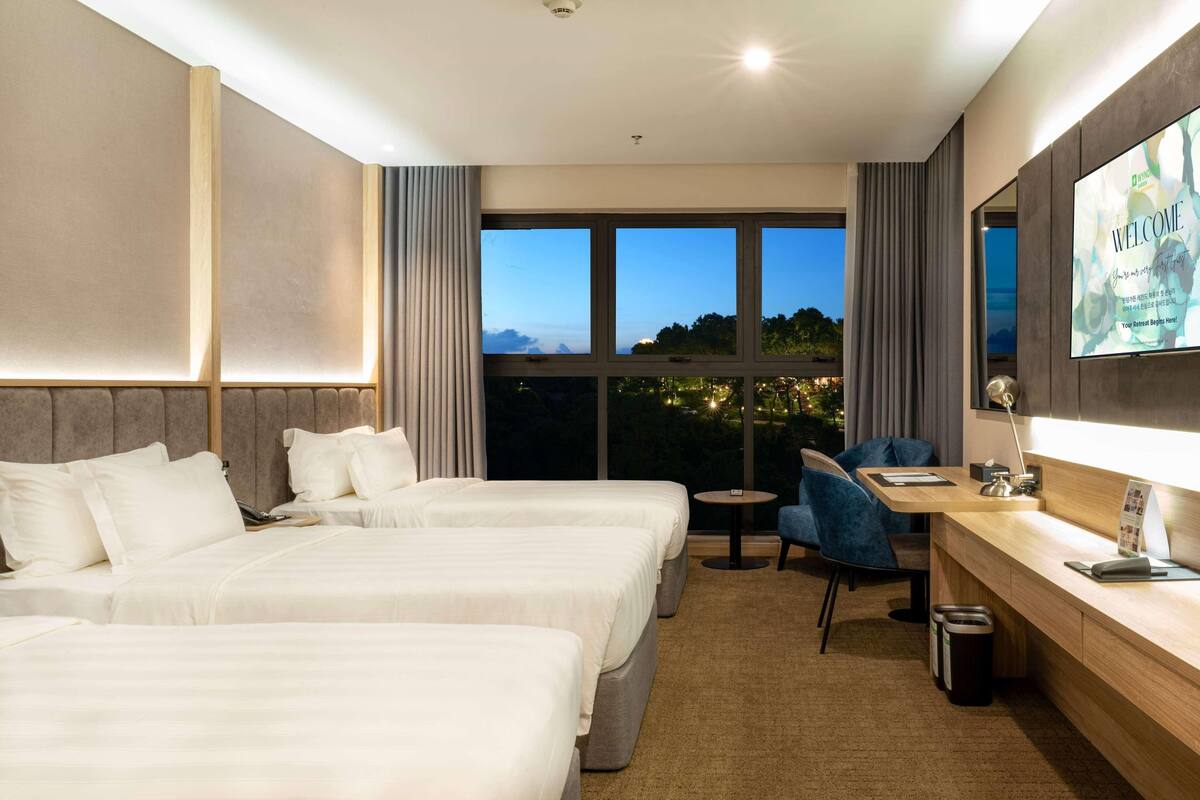 A hotel room with two beds, a desk, a chair, a TV, and a large window with a view.