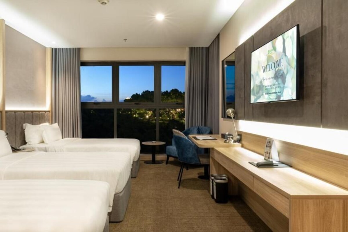 A hotel room with two beds, a desk, a chair, a TV, and large windows with a view of the city at dusk.