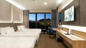A hotel room with two beds, a desk, a chair, a TV, and large windows with a view of the city at dusk. - Wyndham Garden Legend Halong (Bai Chay)