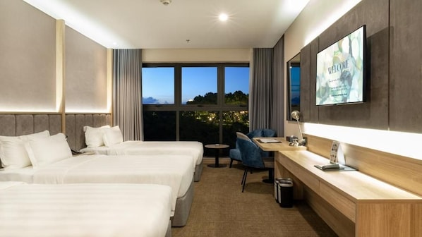 A hotel room with two beds, a desk, a chair, a TV, and large windows with a view of the city at dusk. - Wyndham Garden Legend Halong (Bai Chay)