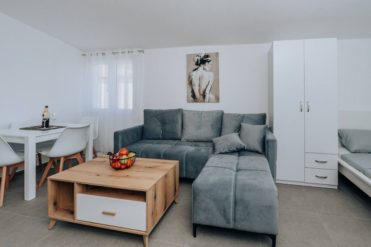A modern living room with a grey sofa, a wooden coffee table with fruit, a white dining table, and a white cabinet.