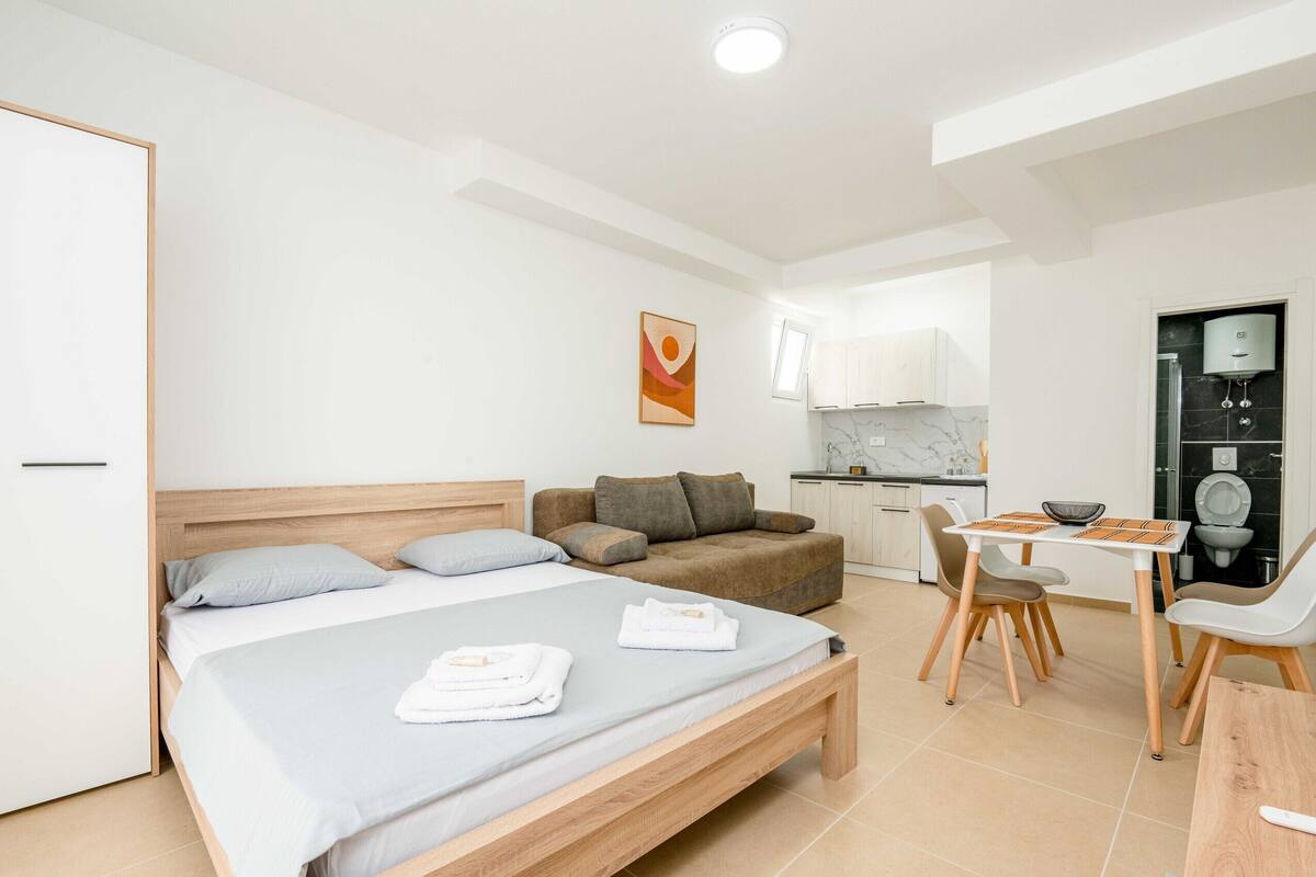 A modern bedroom with a bed, a sofa, a dining table with chairs, and a kitchen area.