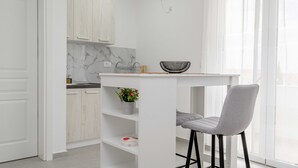Une cuisine moderne avec des meubles blancs, un pare-feu en marbre, un petit îlot central doté d’un récipient et un tabouret de bar.