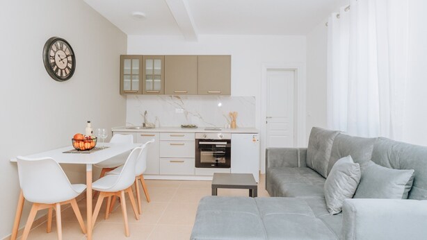 Un salon moderne avec un canapé gris, un coin repas avec des chaises blanches et une cuisine avec des meubles blancs et un four encastré.