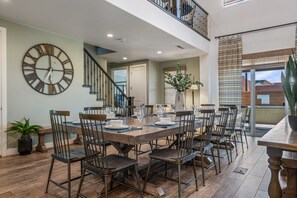 Un comedor amplio con una gran mesa de madera dispuesta para seis personas, rodeada de sillas de metal. Se observa un reloj de pared grande, y una escalera conduce a un nivel superior.