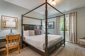 Una cama con dosel de estructura oscura, una silla de madera, una mesita de noche con lámpara y un ventanal con cortinas.
